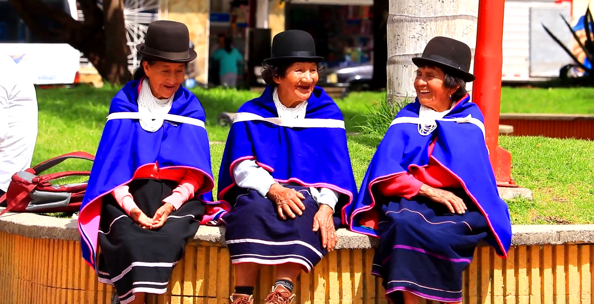 Image shows three women wearing blue indigenous dress sitting together smiling and laughing